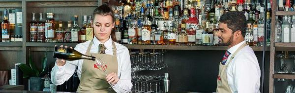 waitress pouring champagne into glass at bar while waiter watches on the side